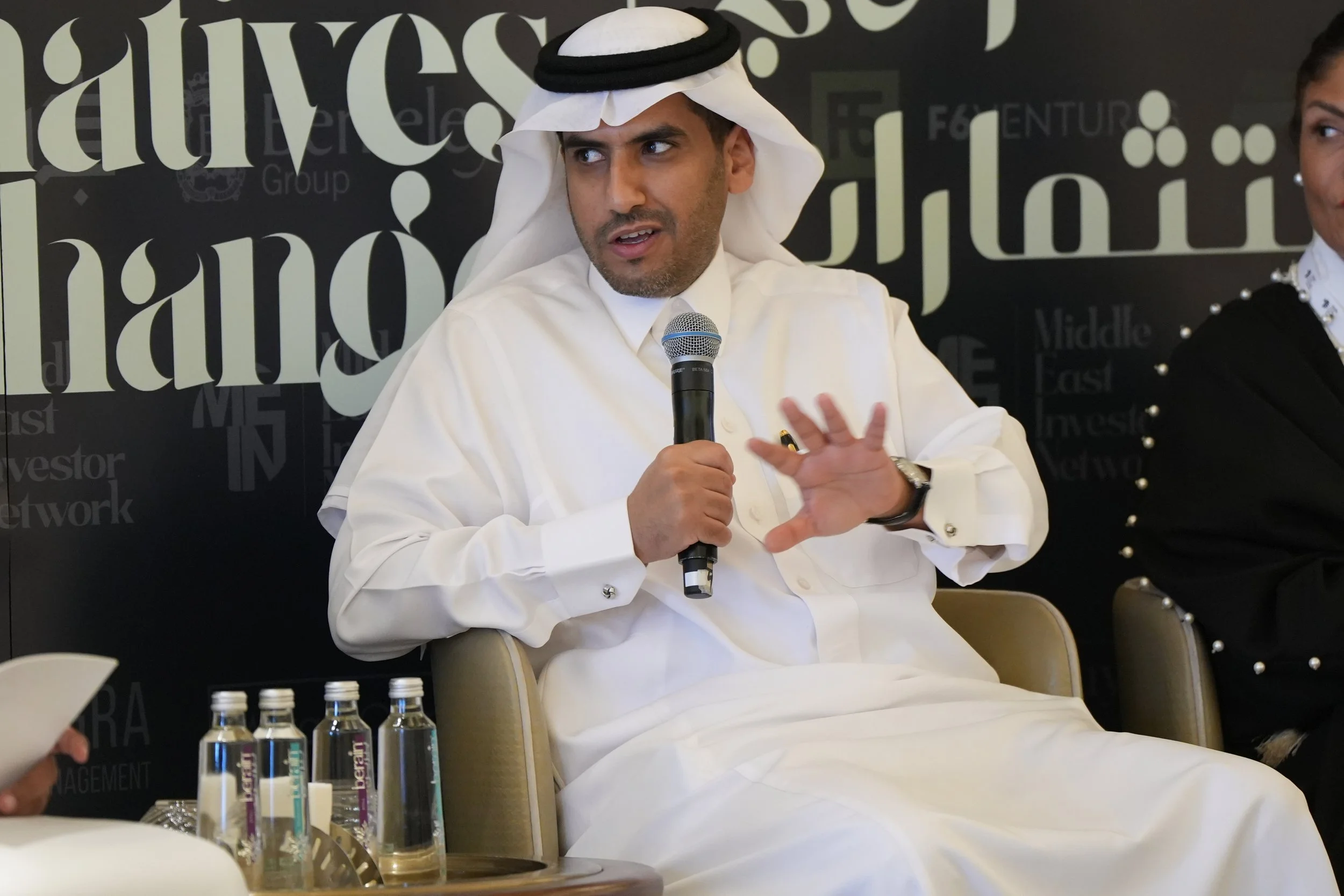 A man wearing traditional Middle Eastern white attire and a black and white patterned headscarf, sitting and speaking into a microphone. Four water bottles are on the table in front of him, and a woman is partially visible on his right. The backgroun