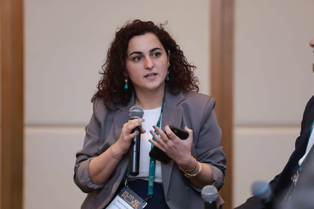 Woman with curly dark hair wearing a gray blazer, turquoise earrings, and has a conference badge around her neck, speaking into a microphone while holding a smartphone.