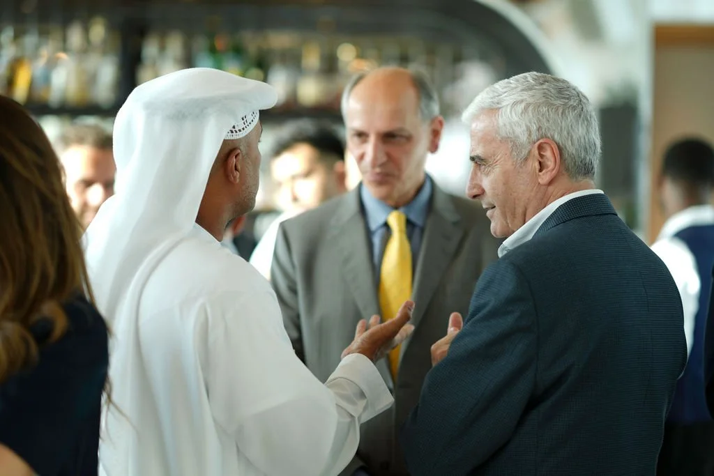 Three men engaged in conversation at a social event, one in traditional Middle Eastern attire, and two in Western business suits, with a bar or restaurant setting in the background.