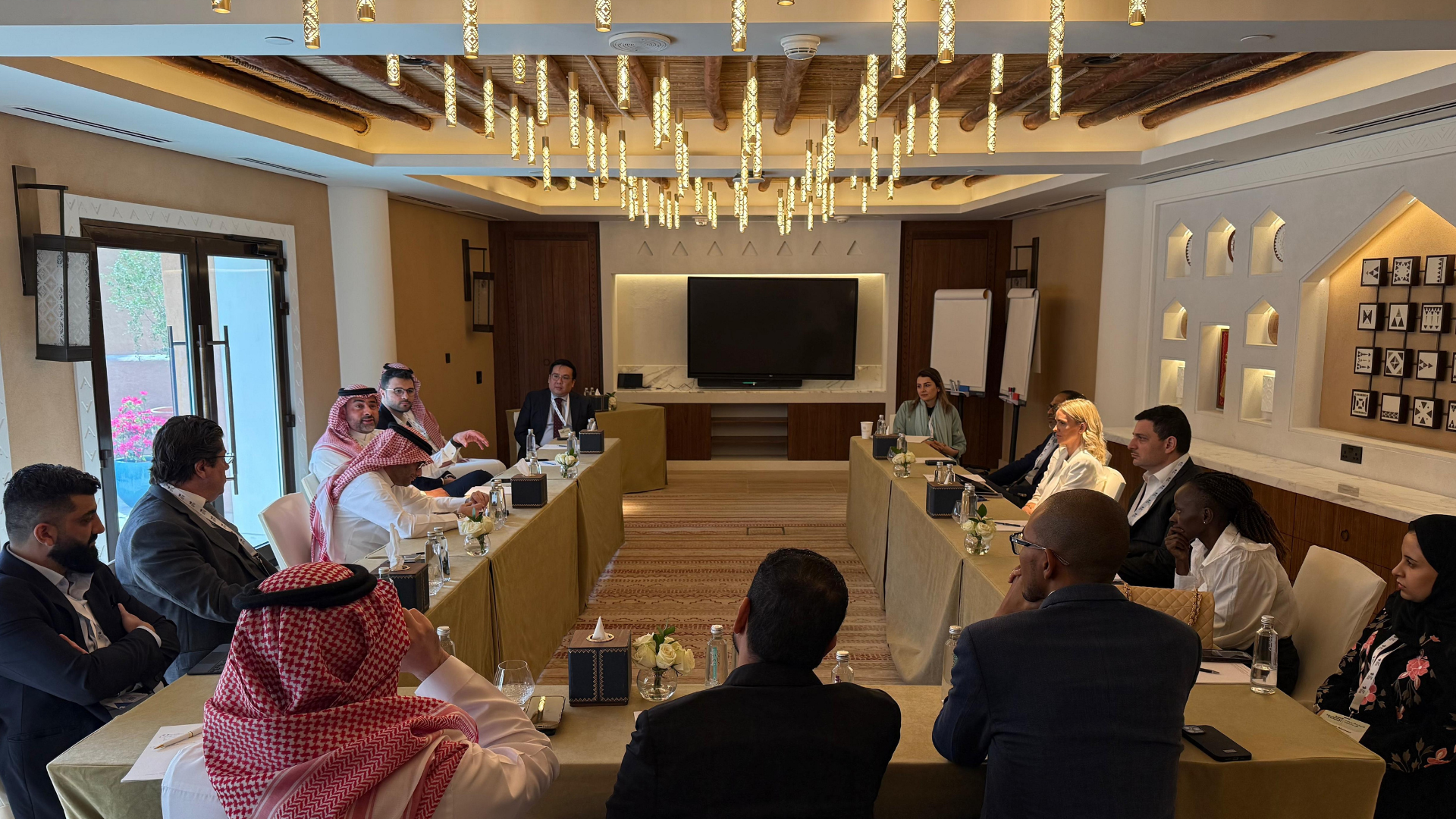 A diverse group of business professionals attending a meeting in a modern conference room. The room is decorated with artistic wall niches, a large television screen, and an elegant hanging light fixture. Participants are seated around tables, listen