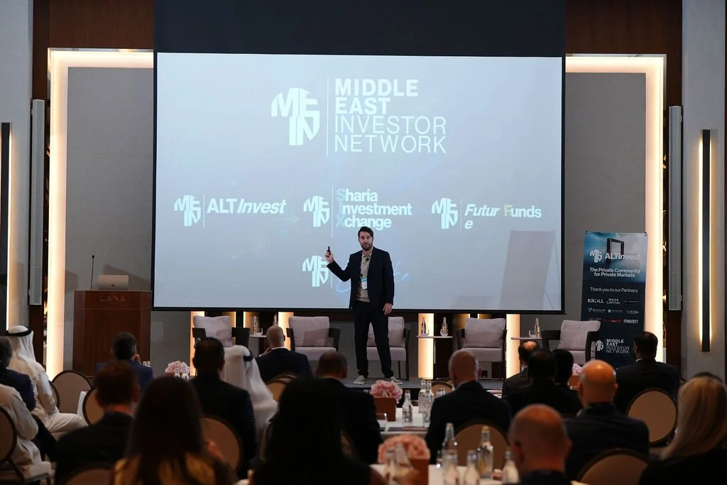 A speaker presenting at the Middle East Investor Network conference, with a large screen displaying the event logo and sponsor names, in a conference room filled with seated attendees.