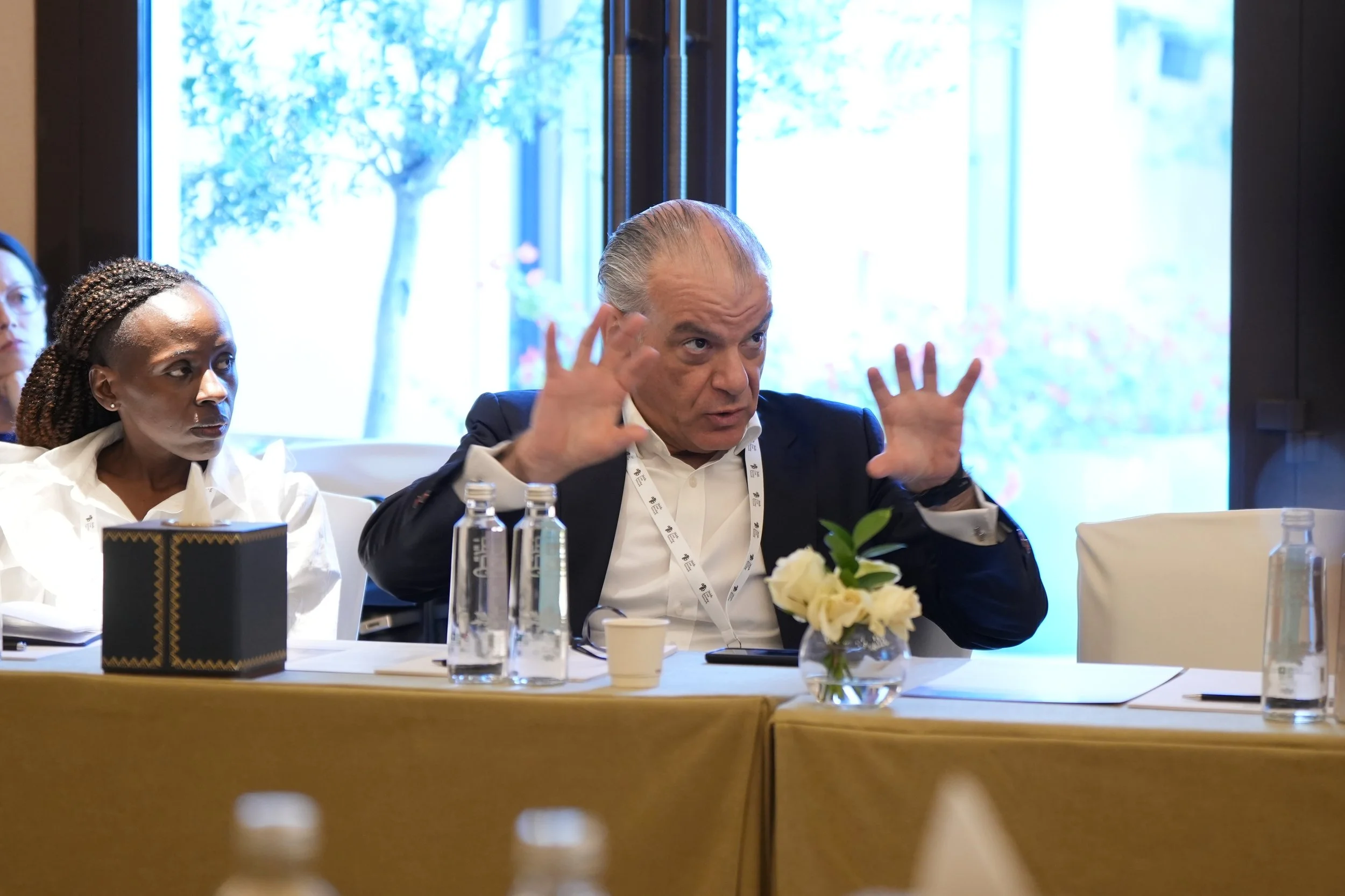 A man in a suit gestures with his hands while sitting at a round table with a woman next to him, during a conference or meeting, with bottles, a flower arrangement, and a laptop on the table.