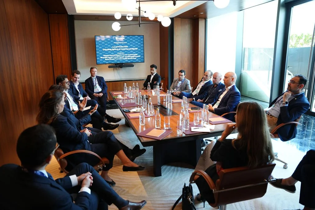 A business meeting in a conference room with 11 people, mostly men, sitting around a large wooden table. There are water bottles, notebooks, and glasses on the table. A flat-screen TV is mounted on the wall displaying a blue screen with white text. T