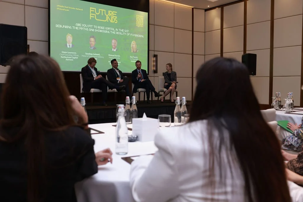 Panel discussion at a conference with four speakers on stage and audience members seated at tables, some taking notes. A large screen behind the panel displays the event's title and topics.
