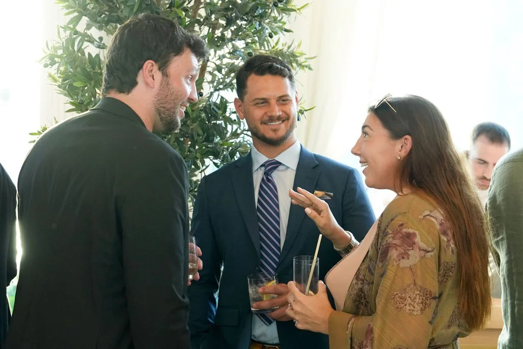 Three people at a social gathering, engaged in conversation, holding drinks, smiling, with a plant and light-colored curtains in the background.