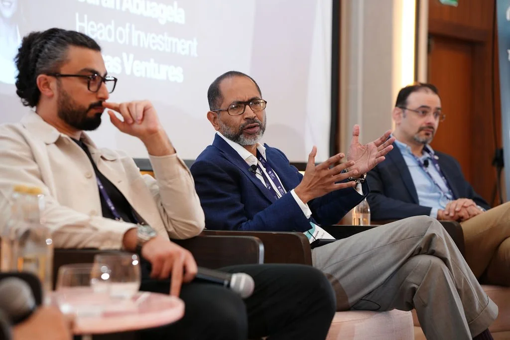 Three men sit on a panel at a conference, with a speaker in the middle actively gesturing while talking. The background features a presentation screen.