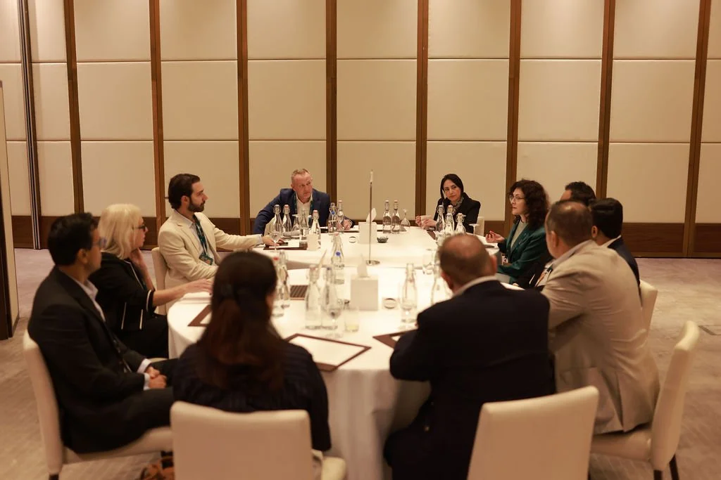 A group of eleven people seated around a rectangular conference table in a professional setting, with notepads, pens, and bottles of water in front of them, engaging in a discussion.
