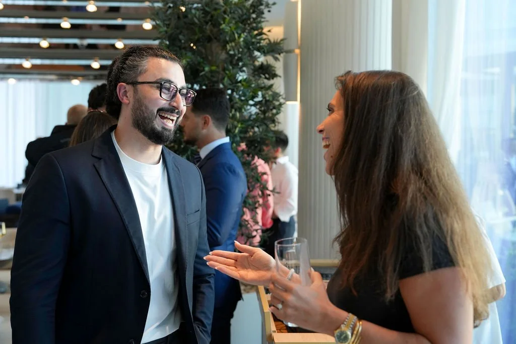 A man with glasses and a beard wearing a dark blazer and white shirt is smiling and talking to a woman with long brown hair, wearing a black top, who is holding a glass of champagne, at a social gathering or party.