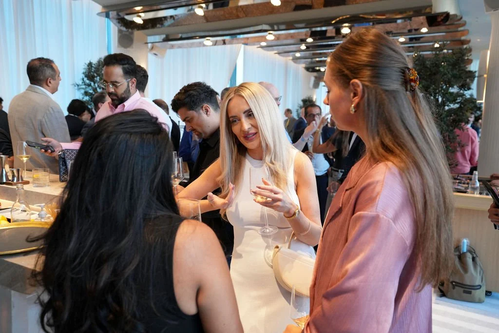 Group of people socializing at a cocktail party, with women engaging in conversation and holding wine glasses, in a well-lit, modern venue.