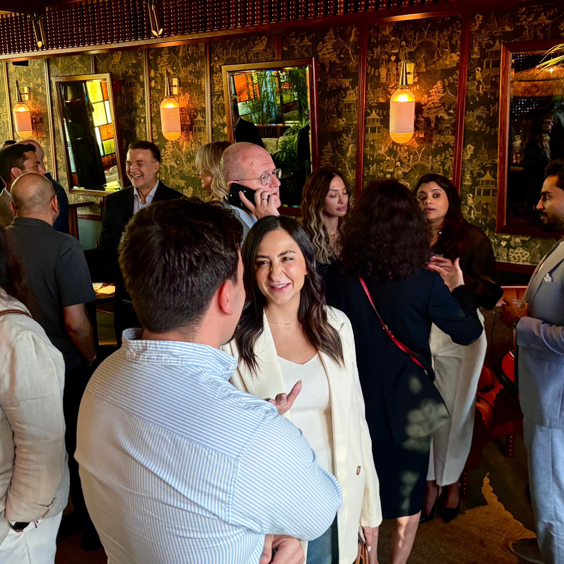 Group of people socializing at a restaurant, with some engaged in conversation and smiling, behind a decorative wall with hanging lanterns.