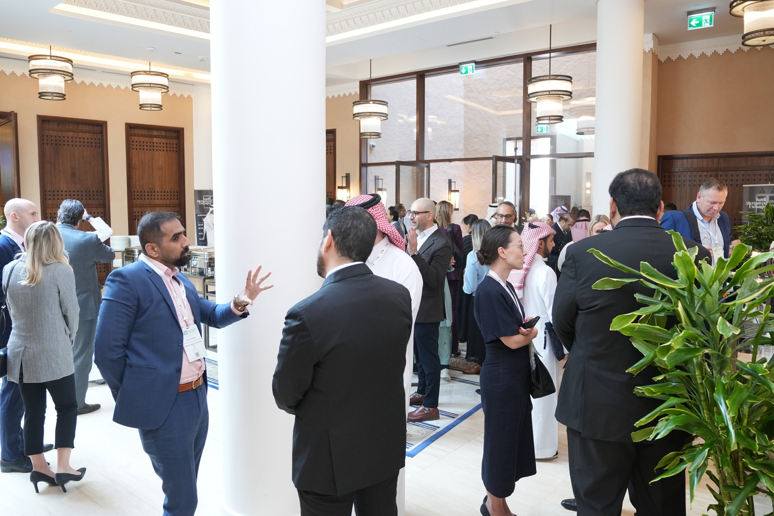 A large group of diverse people dressed in business attire socializing and networking at an indoor conference or event, with some standing in small groups and engaging in conversations, in a well-lit venue with decorative lighting and plants.