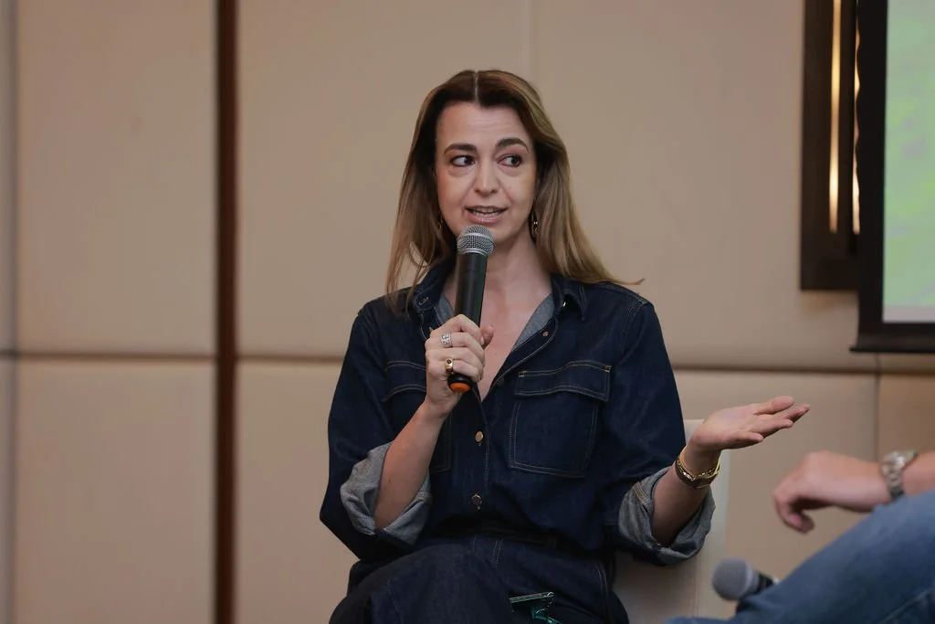 A woman wearing a dark denim shirt speaking into a microphone during a discussion or presentation.