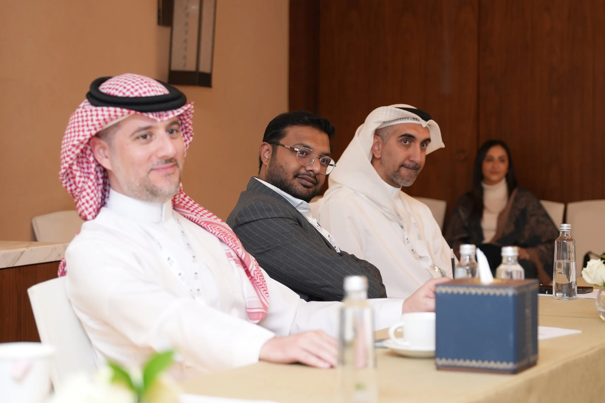 Three men and a woman sitting at a conference table during a meeting; two men are of Middle Eastern descent wearing traditional headdresses, one man appears to be of South Asian descent wearing glasses and a pinstriped suit, the woman in the backgrou