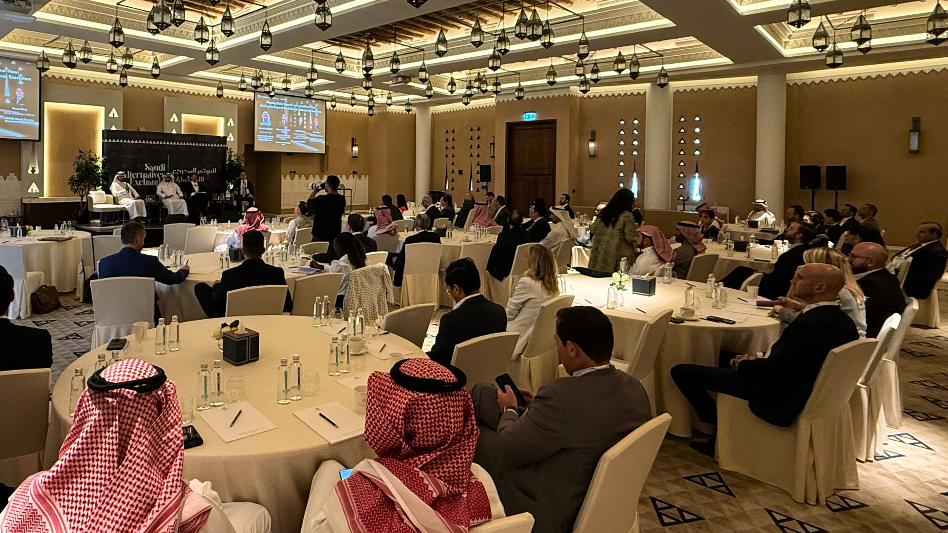 A large conference room filled with attendees sitting at round tables covered with white tablecloths. Several people are dressed in traditional Middle Eastern attire, while others wear business suits. There is a stage at the front with three individu