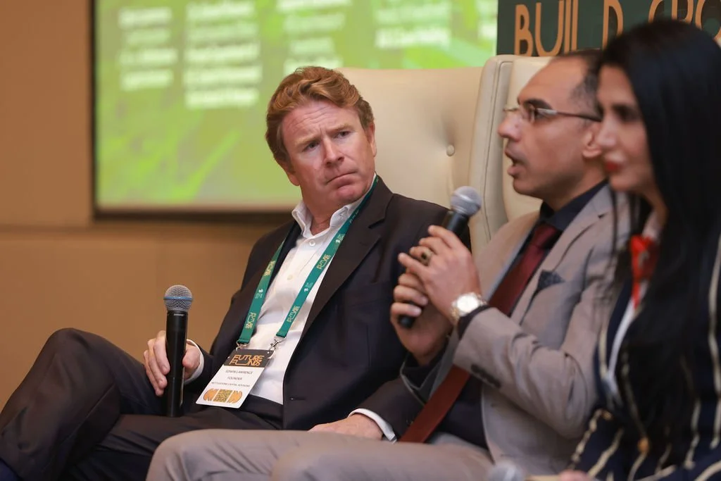 Three individuals seated on a stage, engaged in a panel discussion at a conference, with microphones in hand and a green presentation screen in the background.