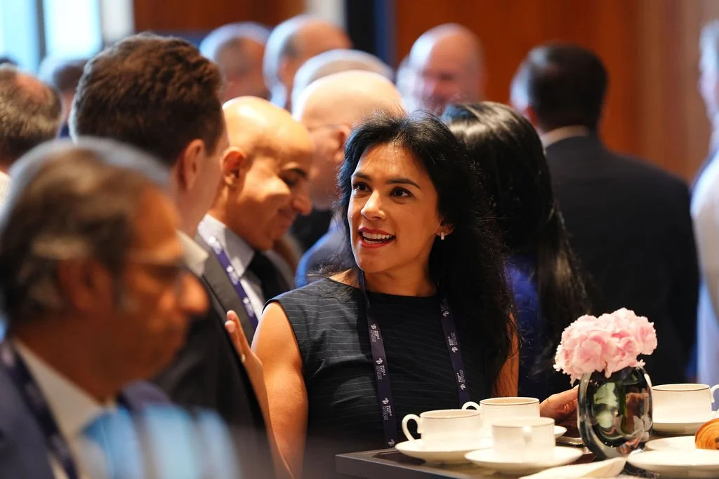 A group of people socializing at a professional event, with a woman in a black dress smiling and talking to others near a table with white cups and a pink flower in a black vase.