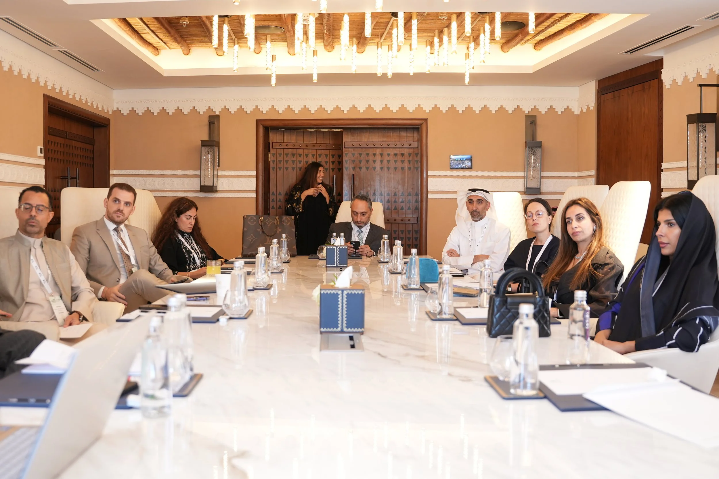 A diverse group of people seated around a large conference table in a well-lit, elegant room with beige walls, wooden accents, and modern chandeliers, engaging in a business meeting.