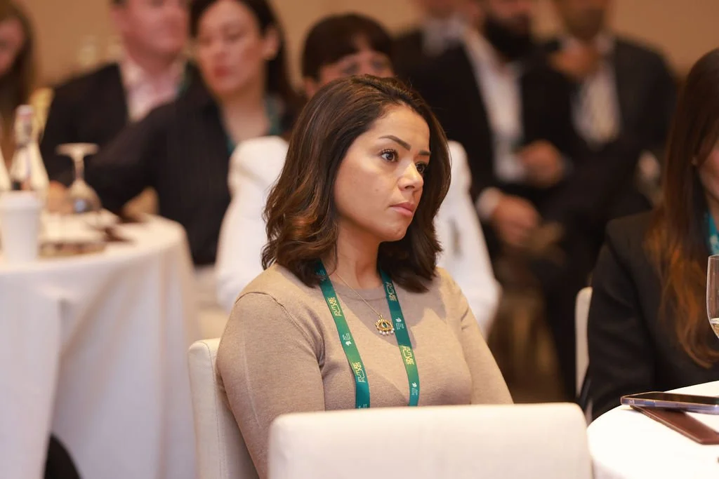 A woman with shoulder-length dark hair attentively listening in a conference room, wearing a beige top and a conference badge.
