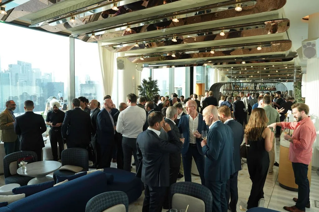 A busy corporate gathering or networking event in a modern high-rise venue with floor-to-ceiling windows overlooking a city skyline. People are dressed in business attire, mingling, talking, and holding drinks, with a bar area in the background.