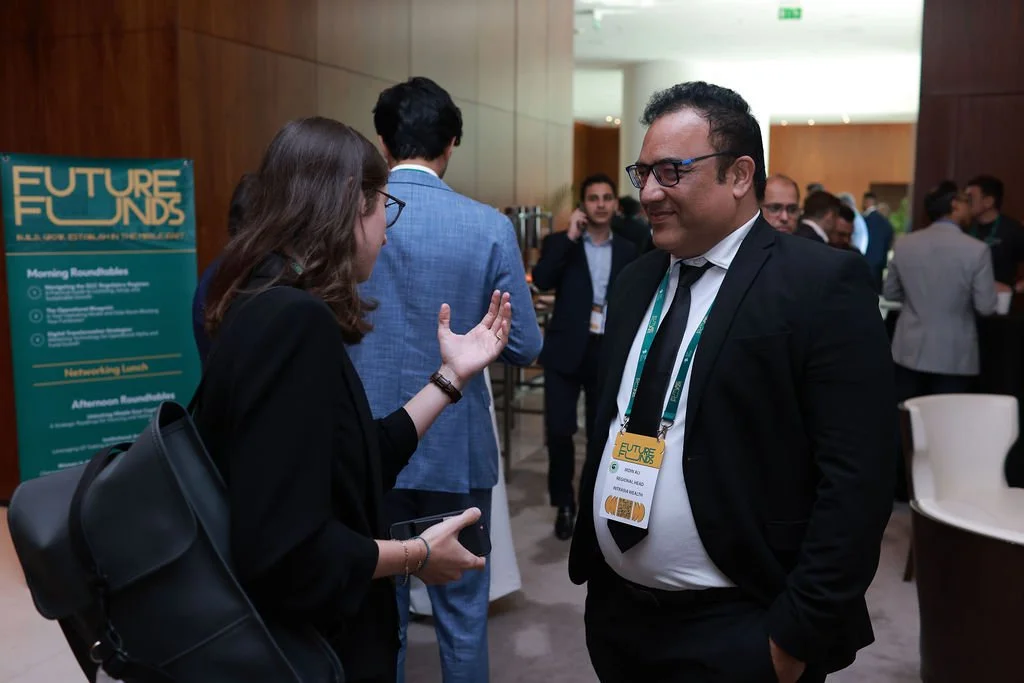 Two people are engaged in conversation at a professional event. The woman has brown hair, glasses, and is wearing a black blazer and carrying a black bag. The man has short dark hair, glasses, and is dressed in a black suit with a white shirt and bla
