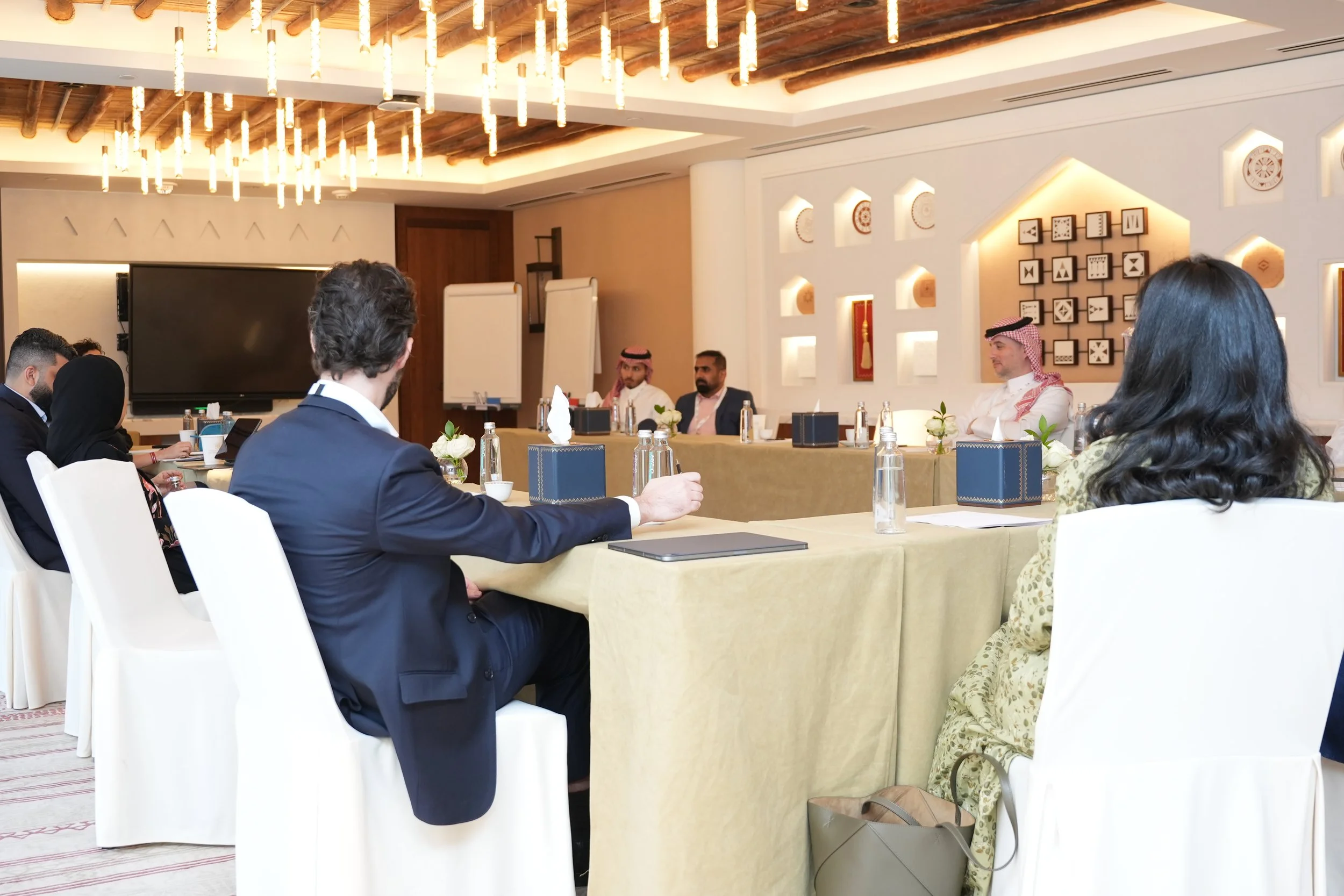 People attending a formal meeting or conference in a room with modern room decor and artwork on the wall, seated around a large table with bottled water, notebooks, and flower arrangements.