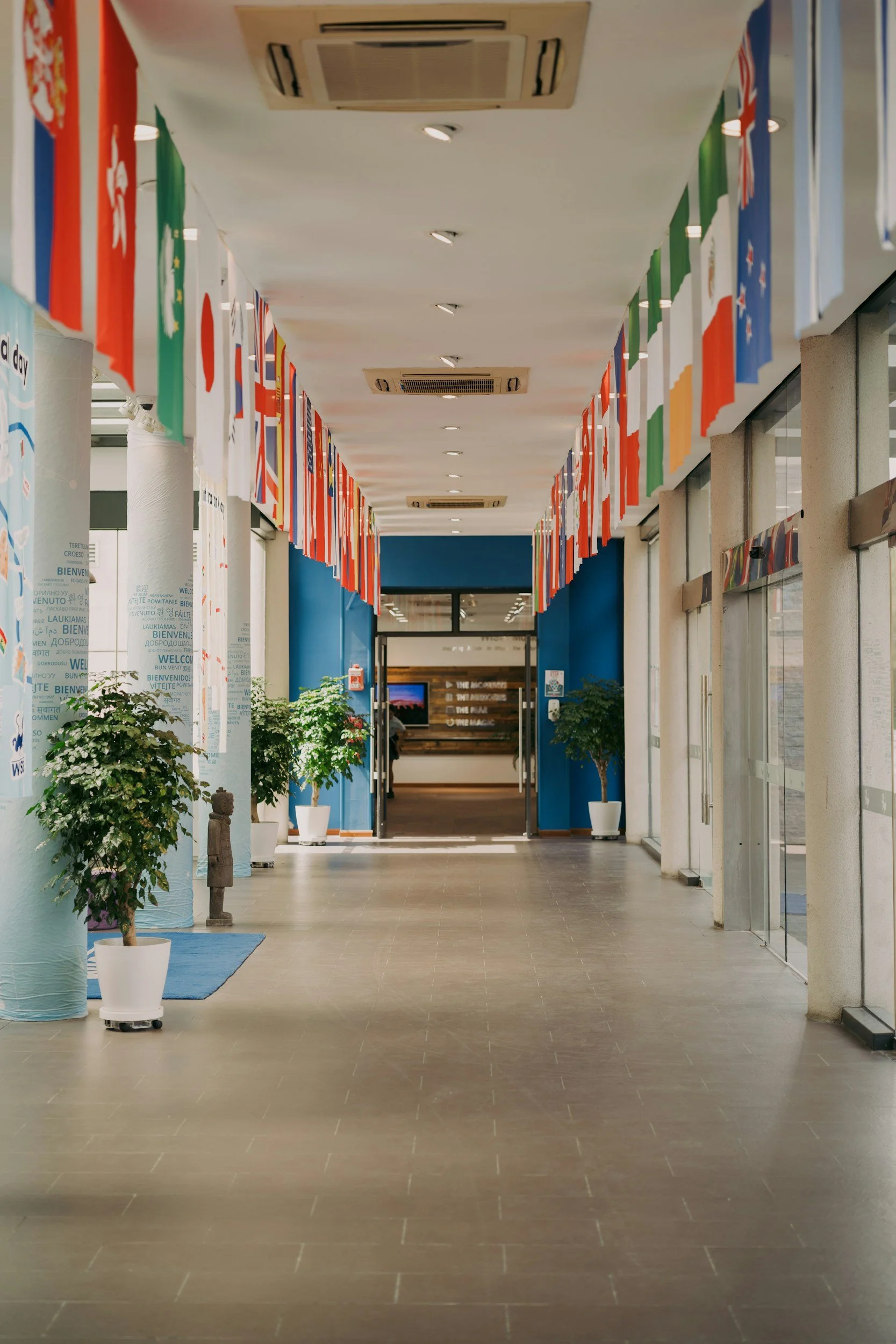 An indoor corridor decorated with international flags hanging from the ceiling, lined with potted plants, with a blue wall at the far end and informational signs.