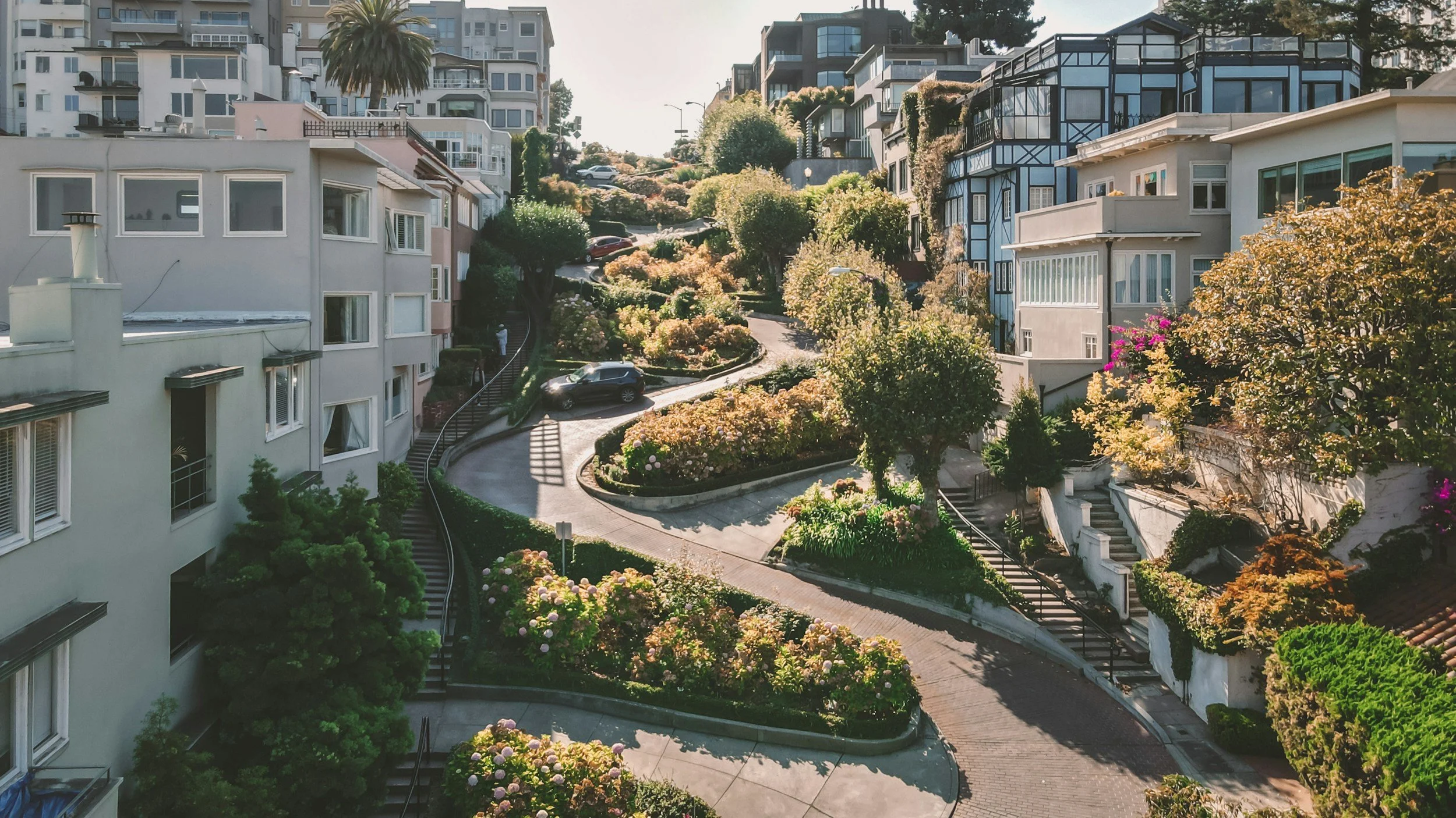 Multiple multi-story apartment or condo buildings with modern architecture, surrounded by lush landscaping and colorful flowering bushes along winding pathways on a hillside in a city.
