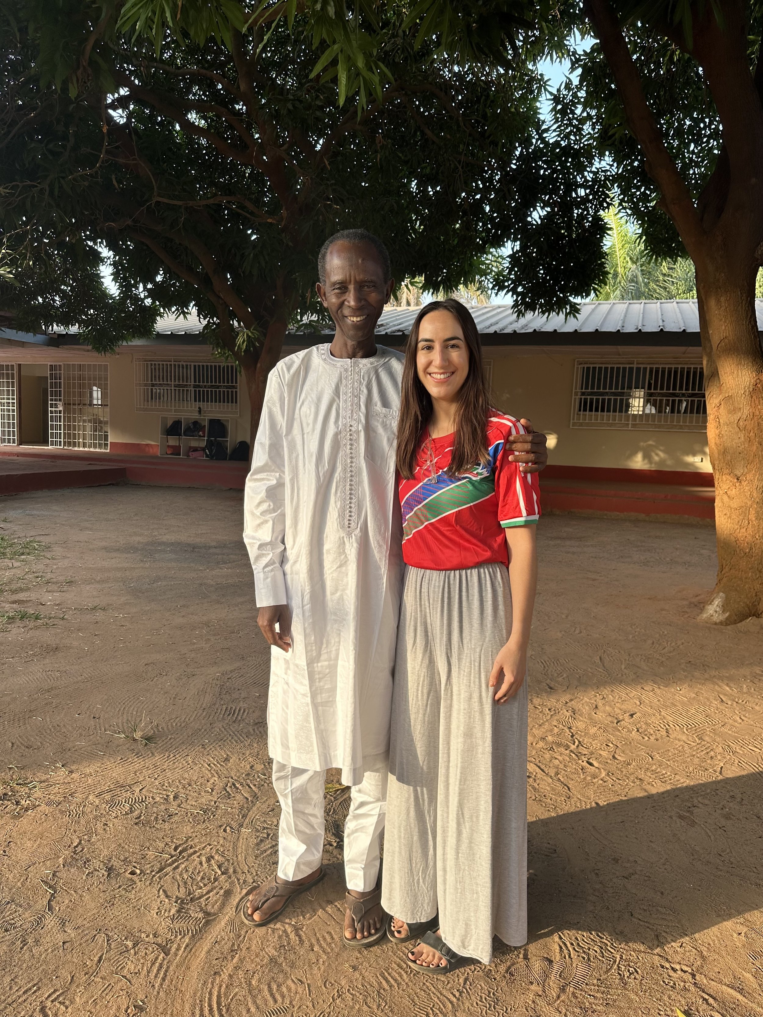 A man and a woman standing outdoors under large trees. The man is wearing a traditional white outfit with sandals, and the woman is wearing a red soccer jersey and beige skirt with sandals. They are smiling and posing for the photo.