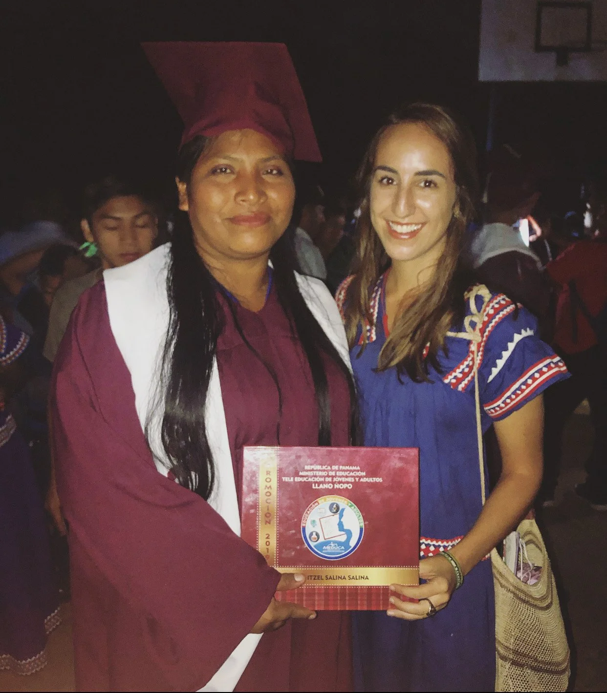 Two women, one in a graduation cap and gown, holding a certificate, smiling at the camera at a graduation ceremony in Latin America. One of the women tutored the other and led professional development for teachers at the international school.