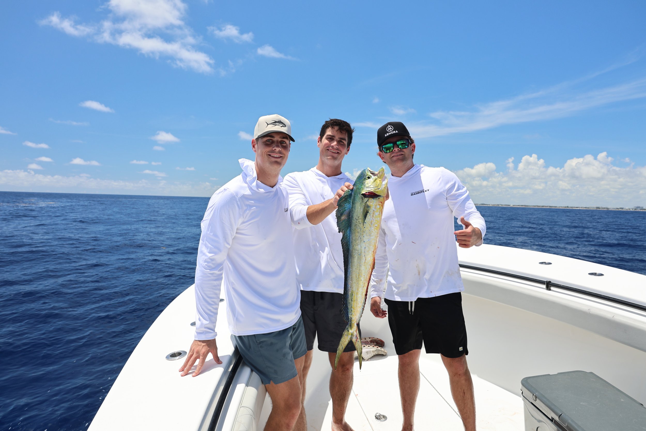 Three men on a boat holding a large fish they caught, with the ocean and a blue sky with clouds in the background.