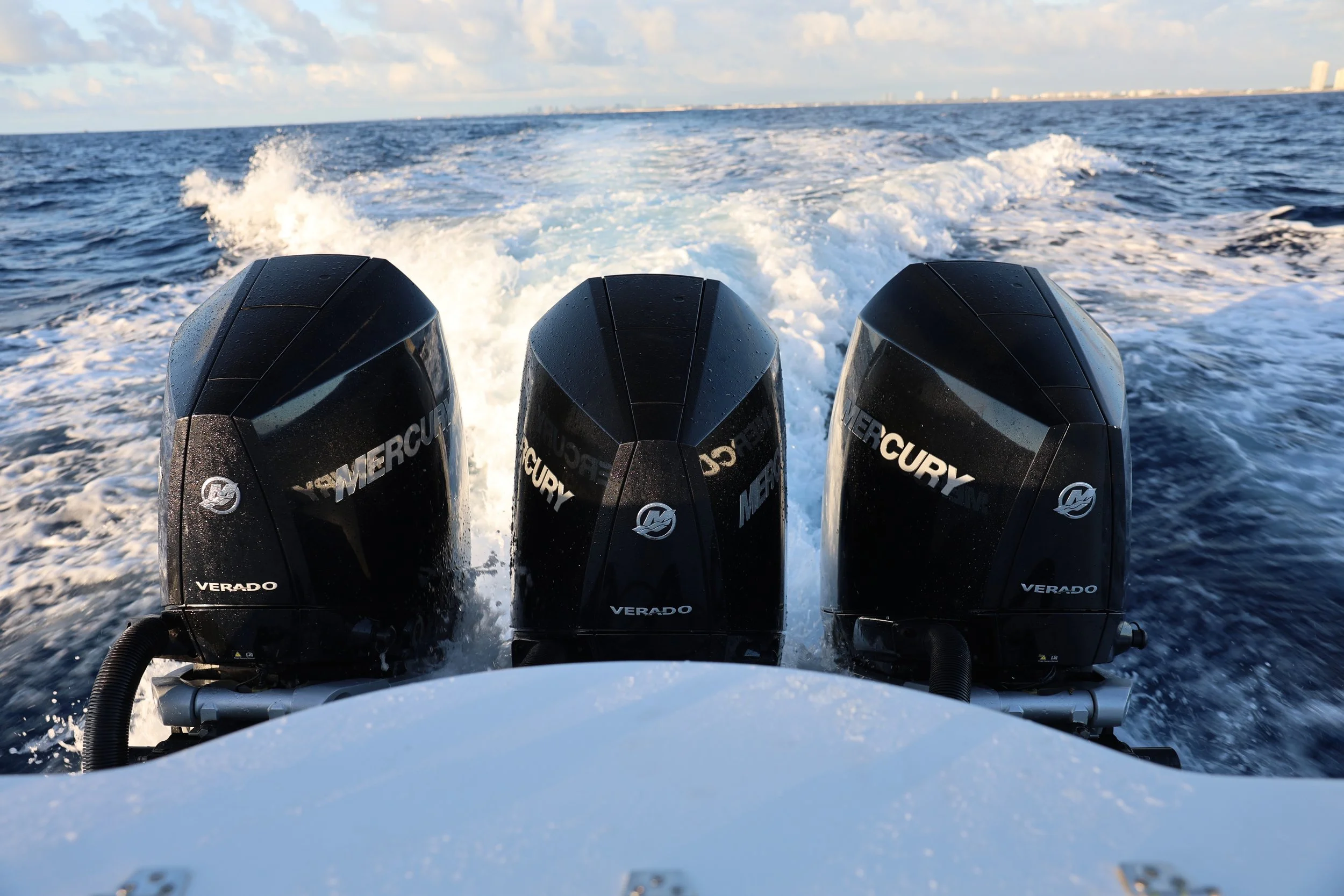Three Mercury Verado outboard motors on a boat engine, with ocean water and city skyline in the distance.