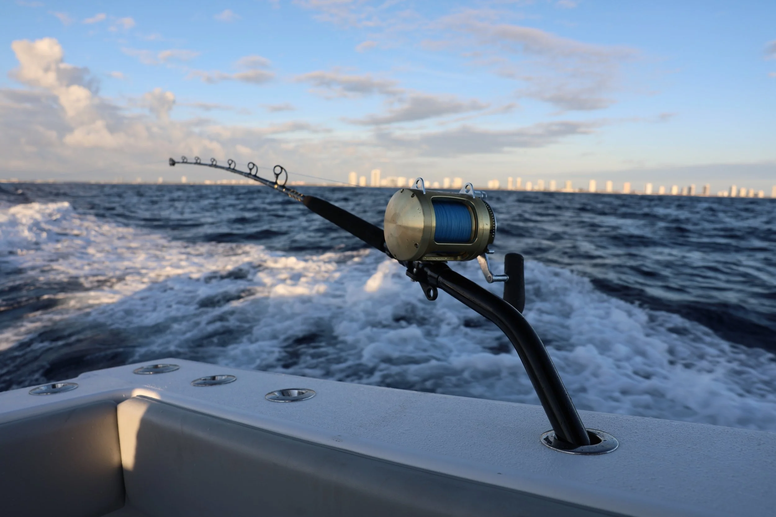 Fishing rod with blue line mounted on a boat, out on a body of water, with a city skyline in the distance under a partly cloudy sky.