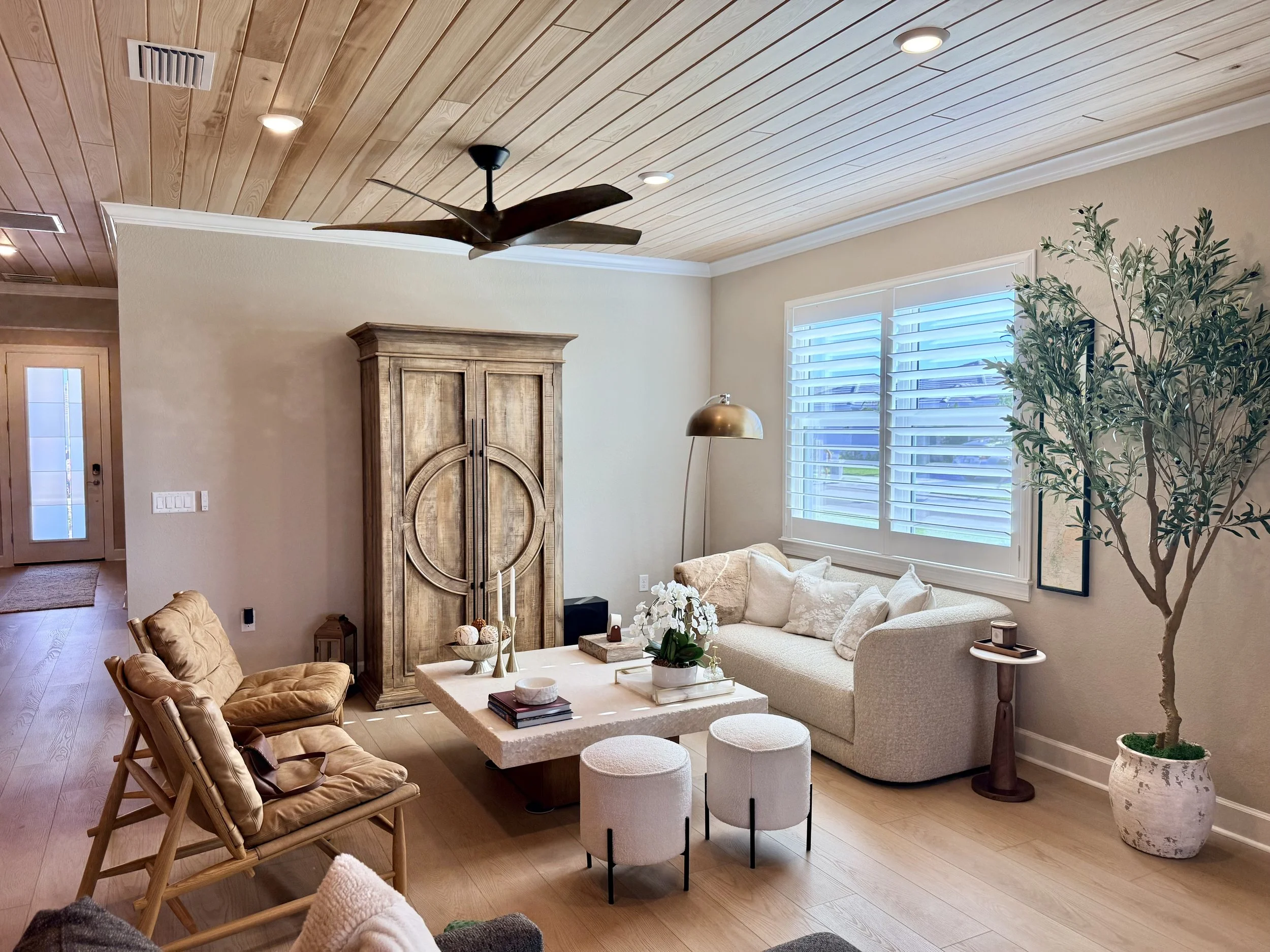 Living room with wood-paneled ceiling, beige walls, large window with white blinds, beige sectional sofa, wooden armchairs with plush cushions, coffee table with decorative items, indoor plant in a large pot, floor lamp, and ceiling fans.