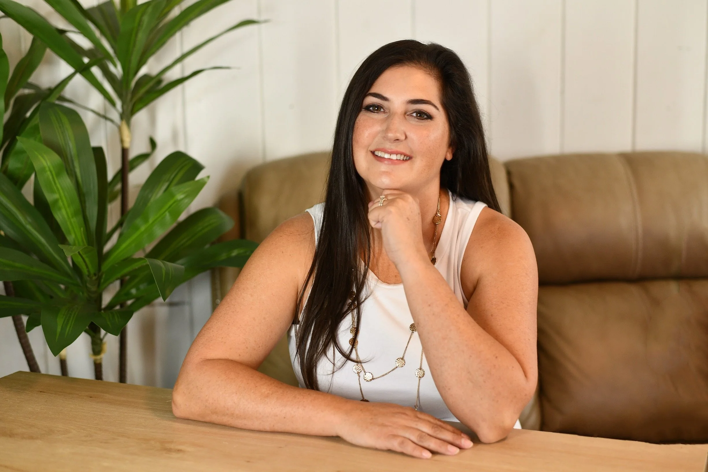 A woman with long brown hair and freckles smiling, sitting at a wooden table with her right arm resting on the table, wearing a white sleeveless top and layered necklaces, with a large green houseplant and beige couch in the background.
