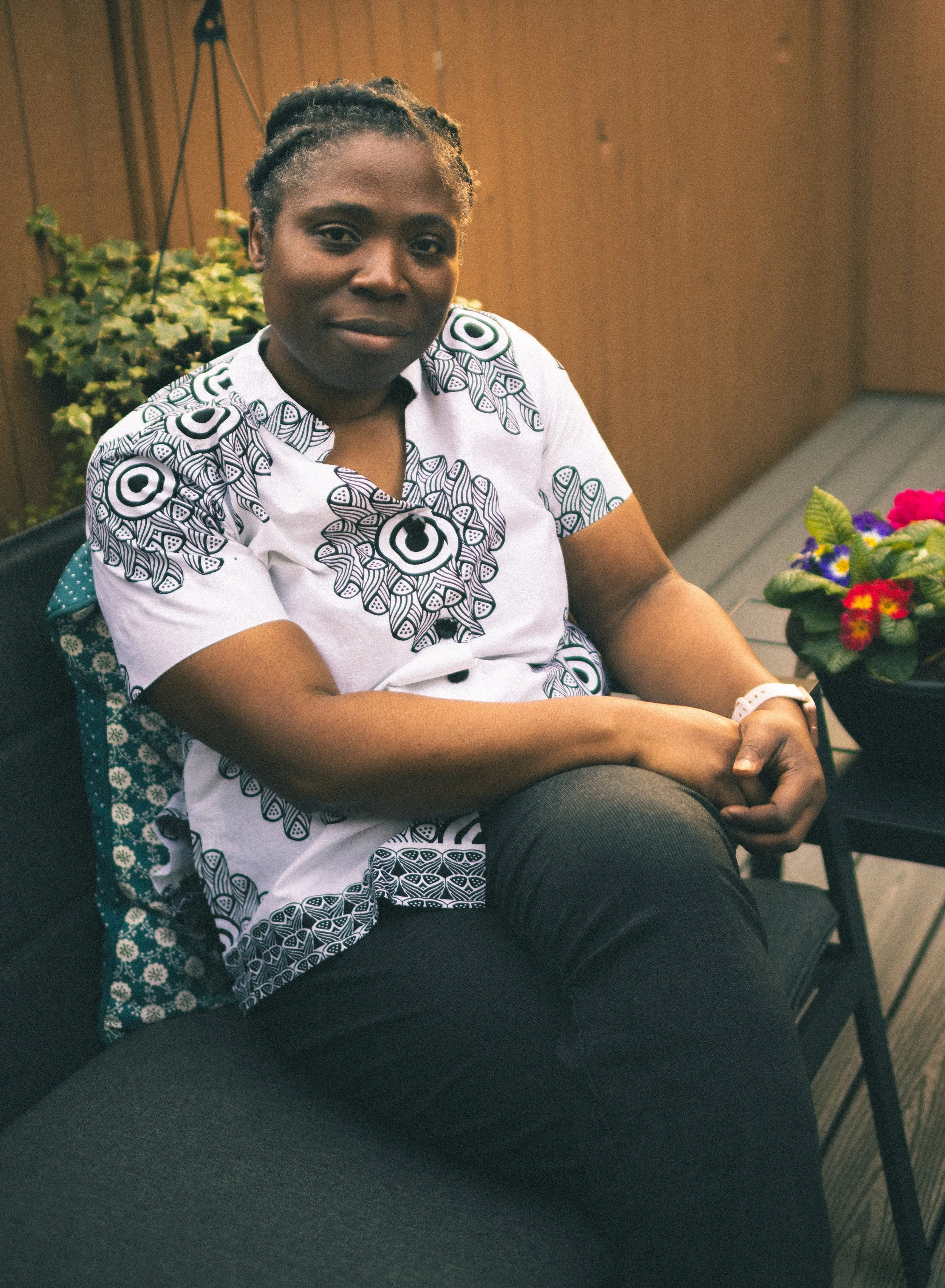 A woman with dark skin, braided hair, wearing a white shirt with black circular patterns, sitting indoors with wooden wall and potted plant in the background, and a colorful flower arrangement on the table in front of her.