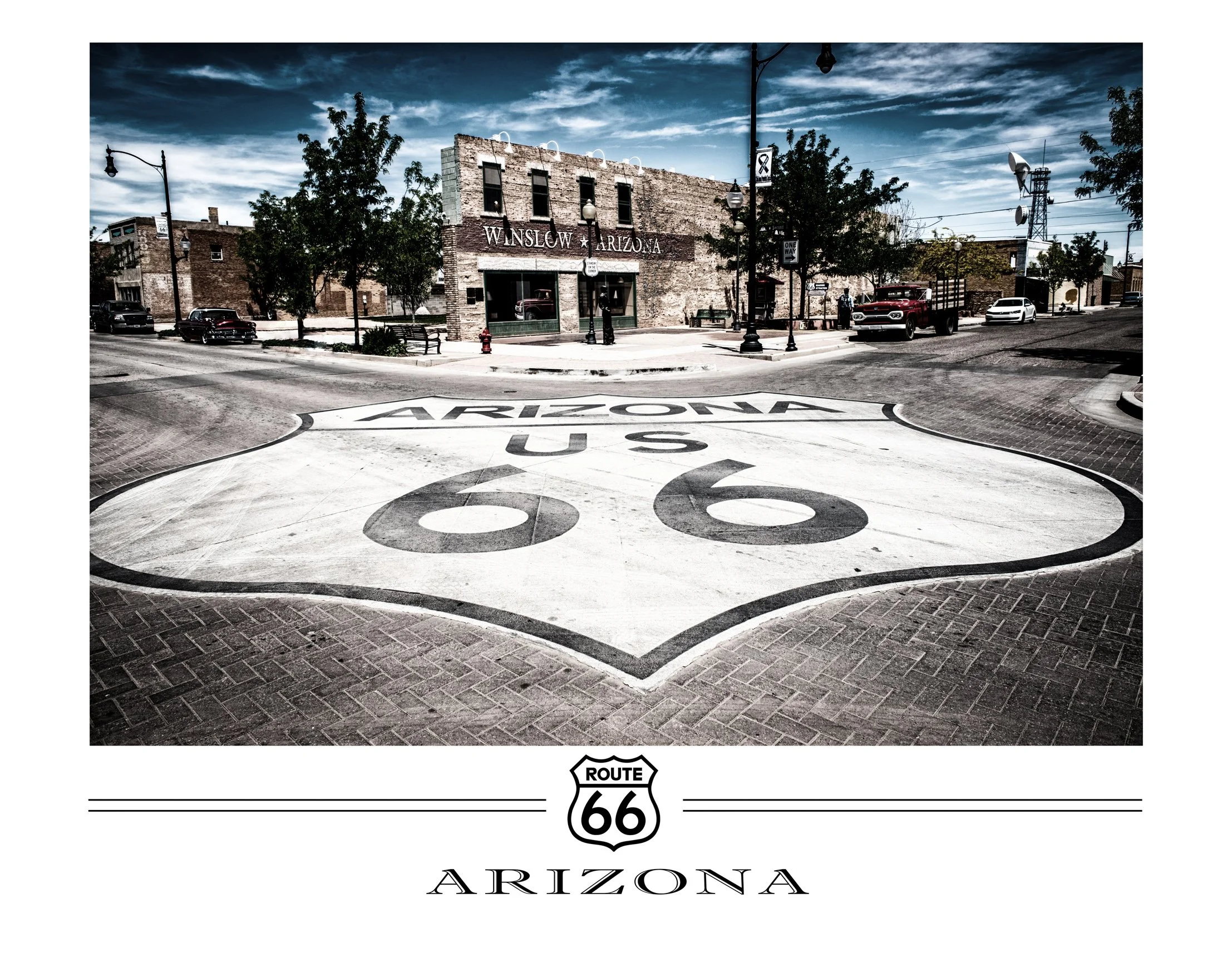 Street view of Winslow, Arizona, with a large Route 66 emblem painted on the ground and historic brick buildings, trees, and cars parked along the street.