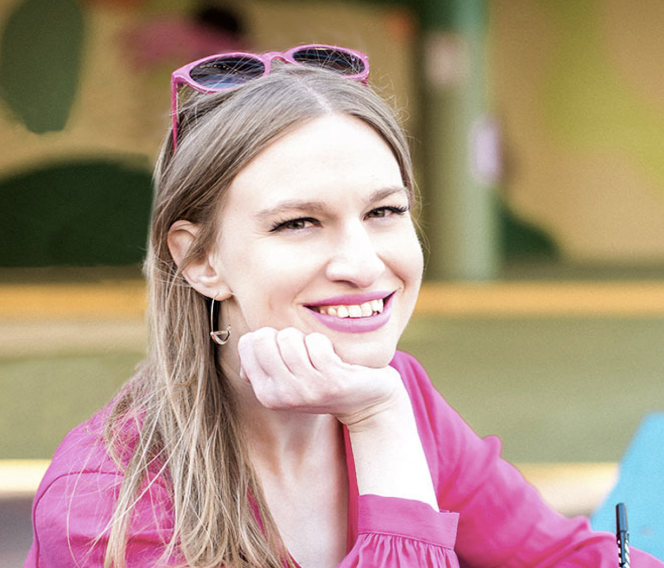 Close-up of a young woman with long hair, wearing pink sunglasses on her head and pink lipstick, smiling outdoors.
