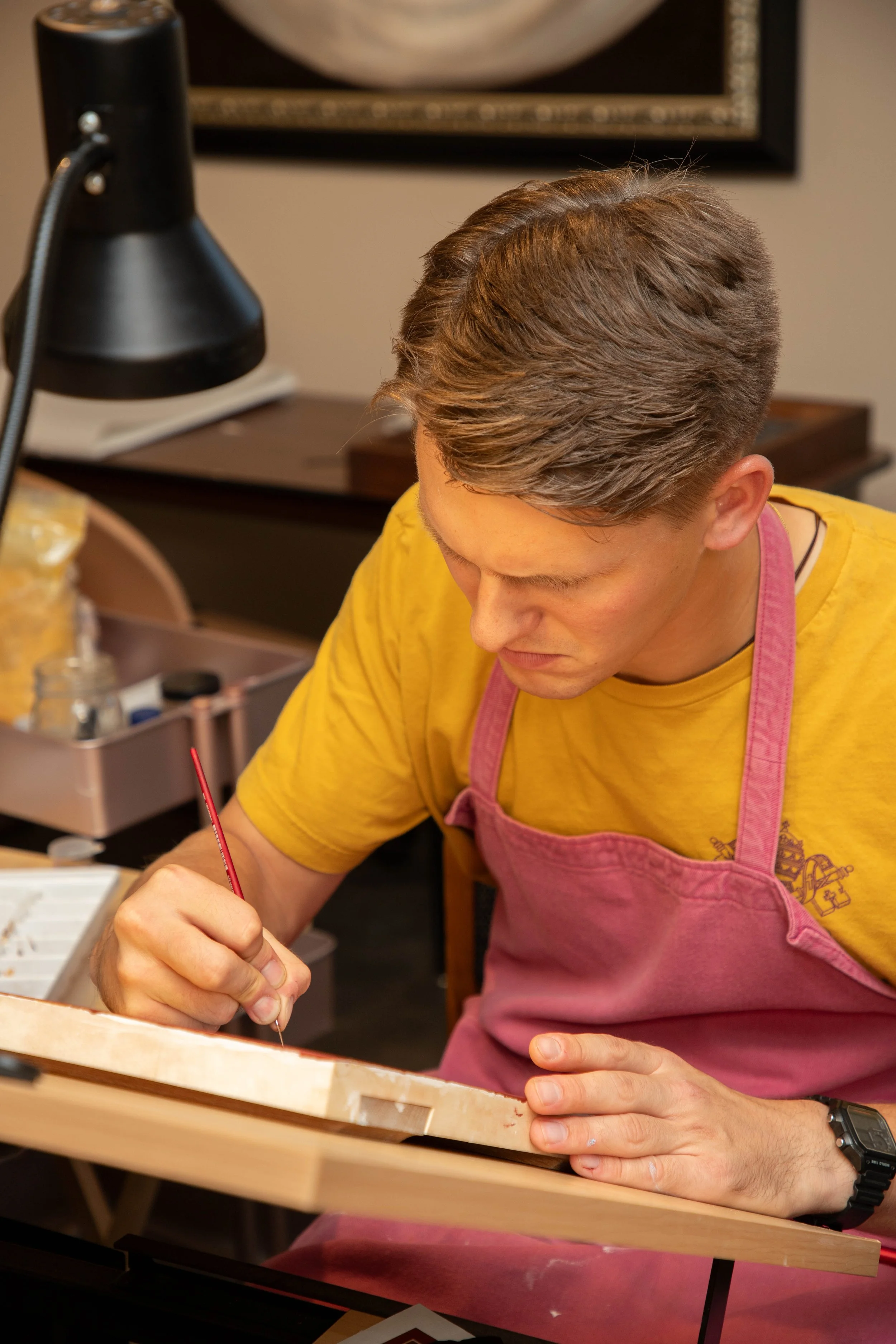Young man in a yellow shirt and pink apron painting a wooden object in an art studio or workshop.