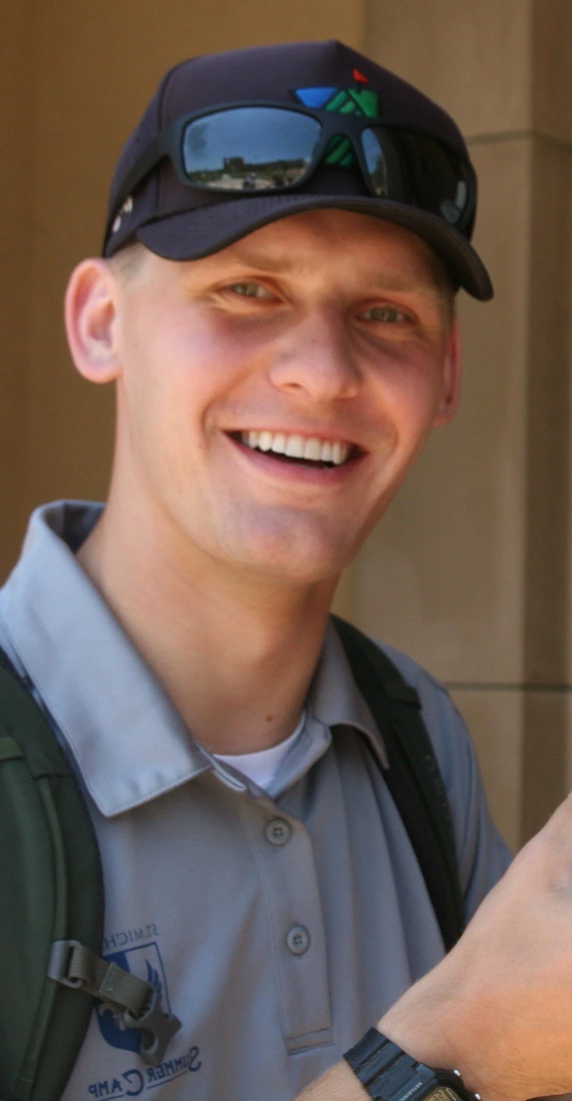A smiling young man wearing glasses on his hat, a backpack, and a gray shirt, standing outdoors.