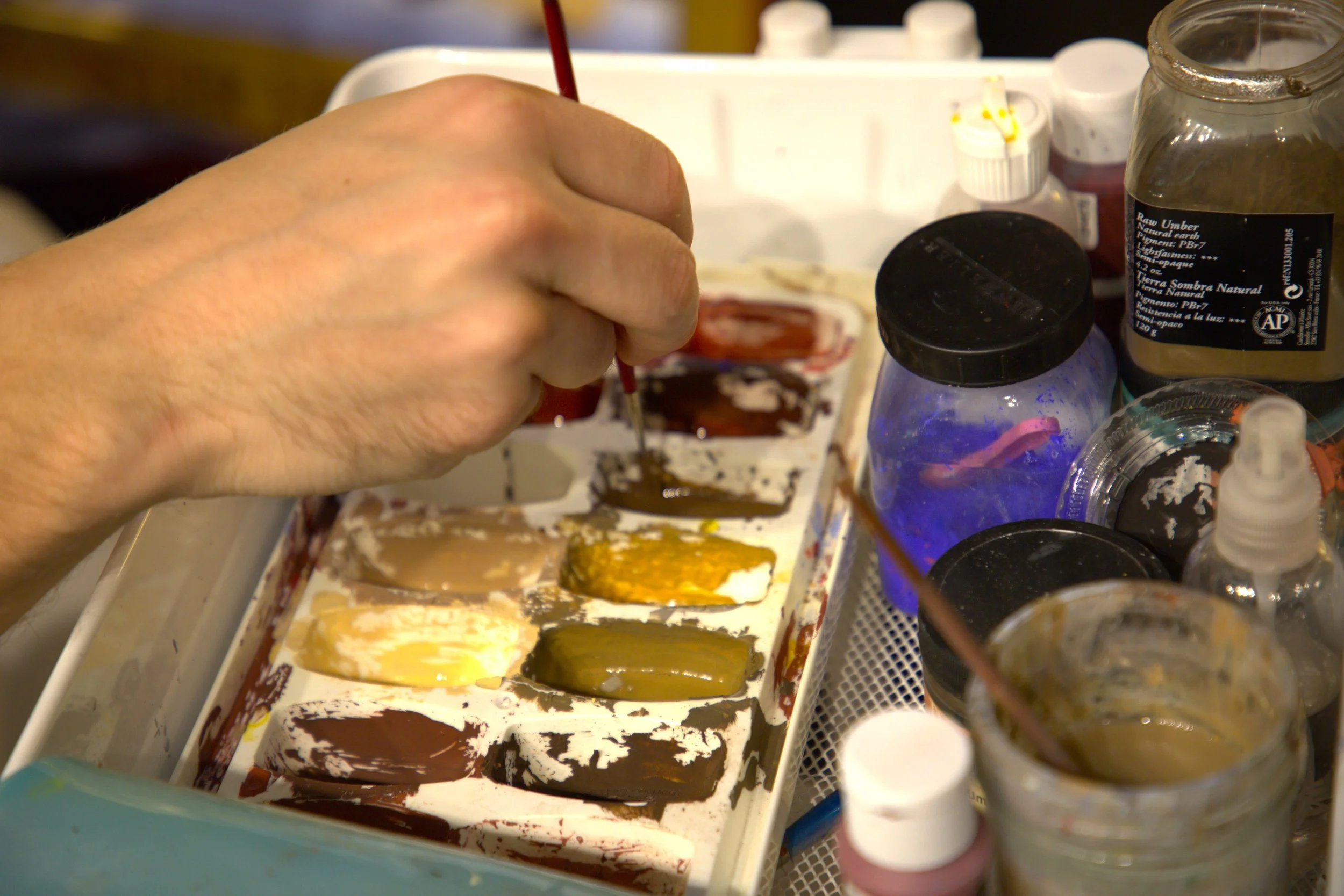 Jake Rogalski is painting with a palette of various colors of egg tempera, with art supplies and jars around.