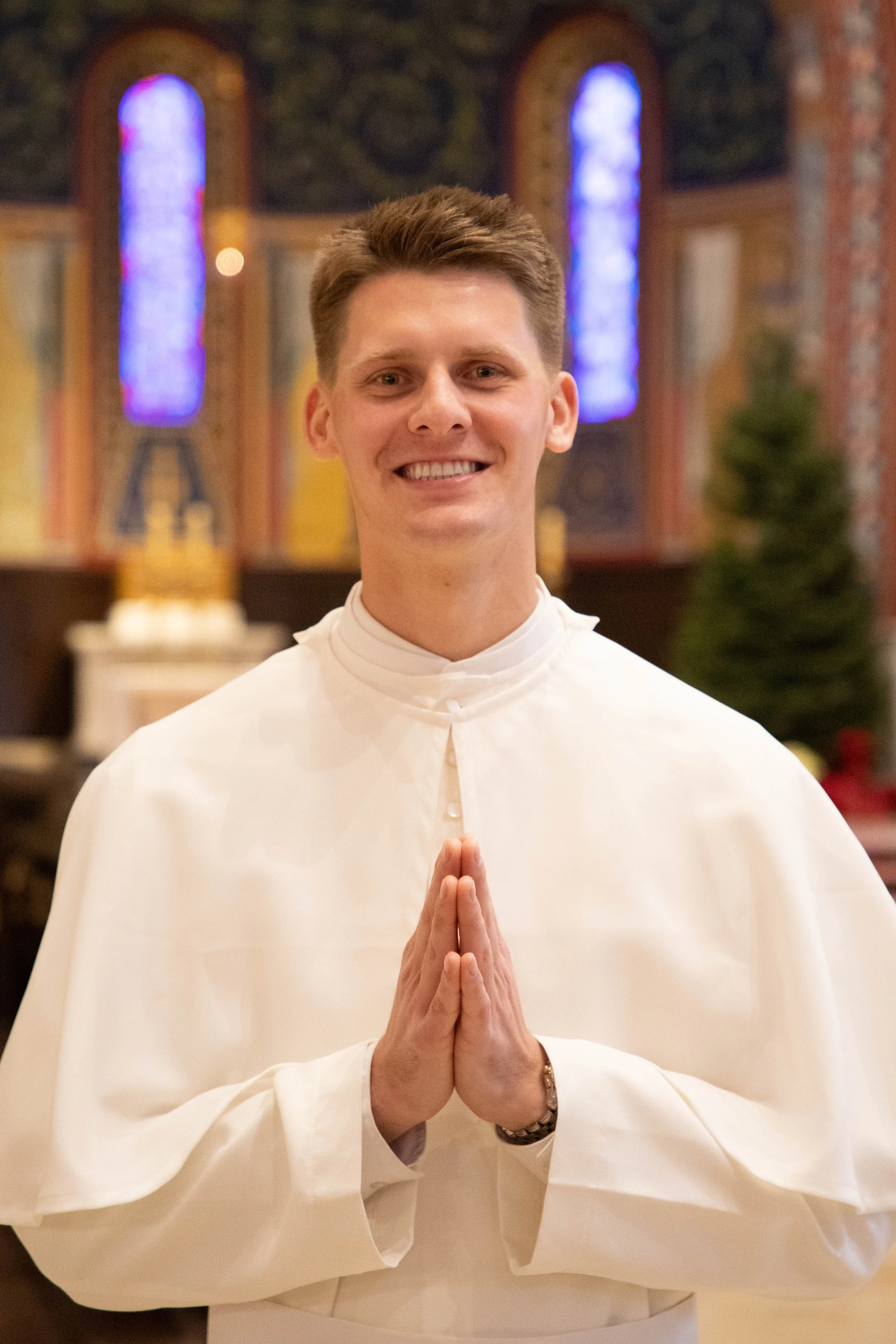 Young man in white clerical robe with hands in prayer position, smiling in church with stained glass windows and ornate decor in the background.