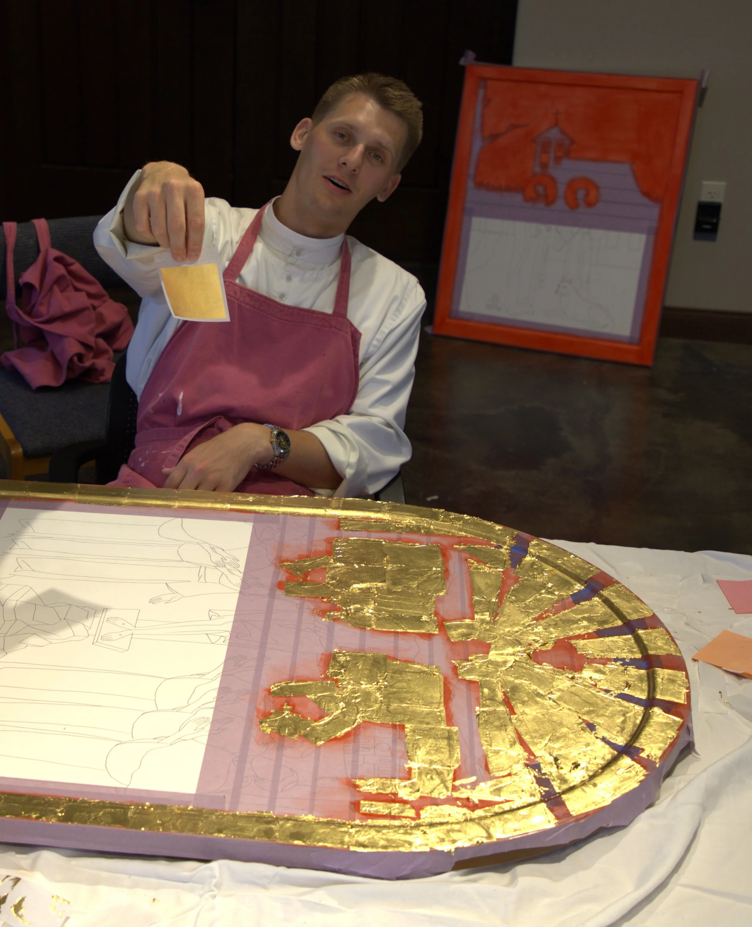Jake Rogalski  holding a small small sheet of gold leaf. There is an icon with gold leaf on it in front of him, and a drawing of a church on an easel in the background.