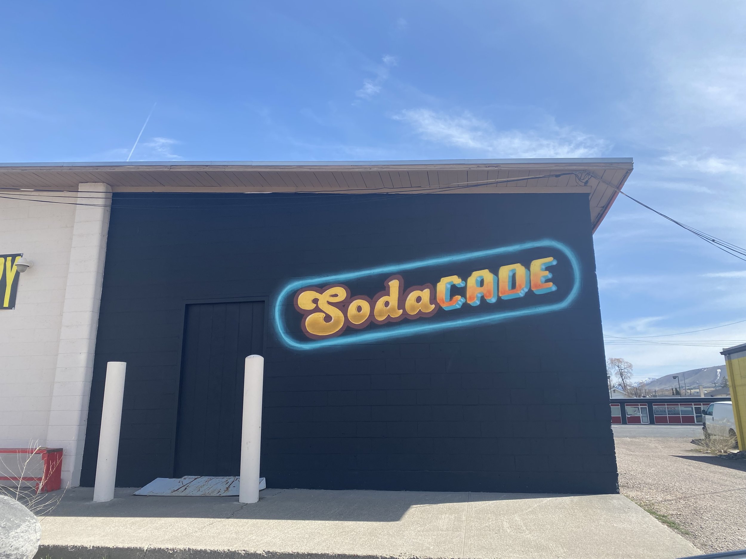 The exterior of a building with a sign that reads 'SodaCade' in colorful neon lights, under a clear blue sky with some clouds.