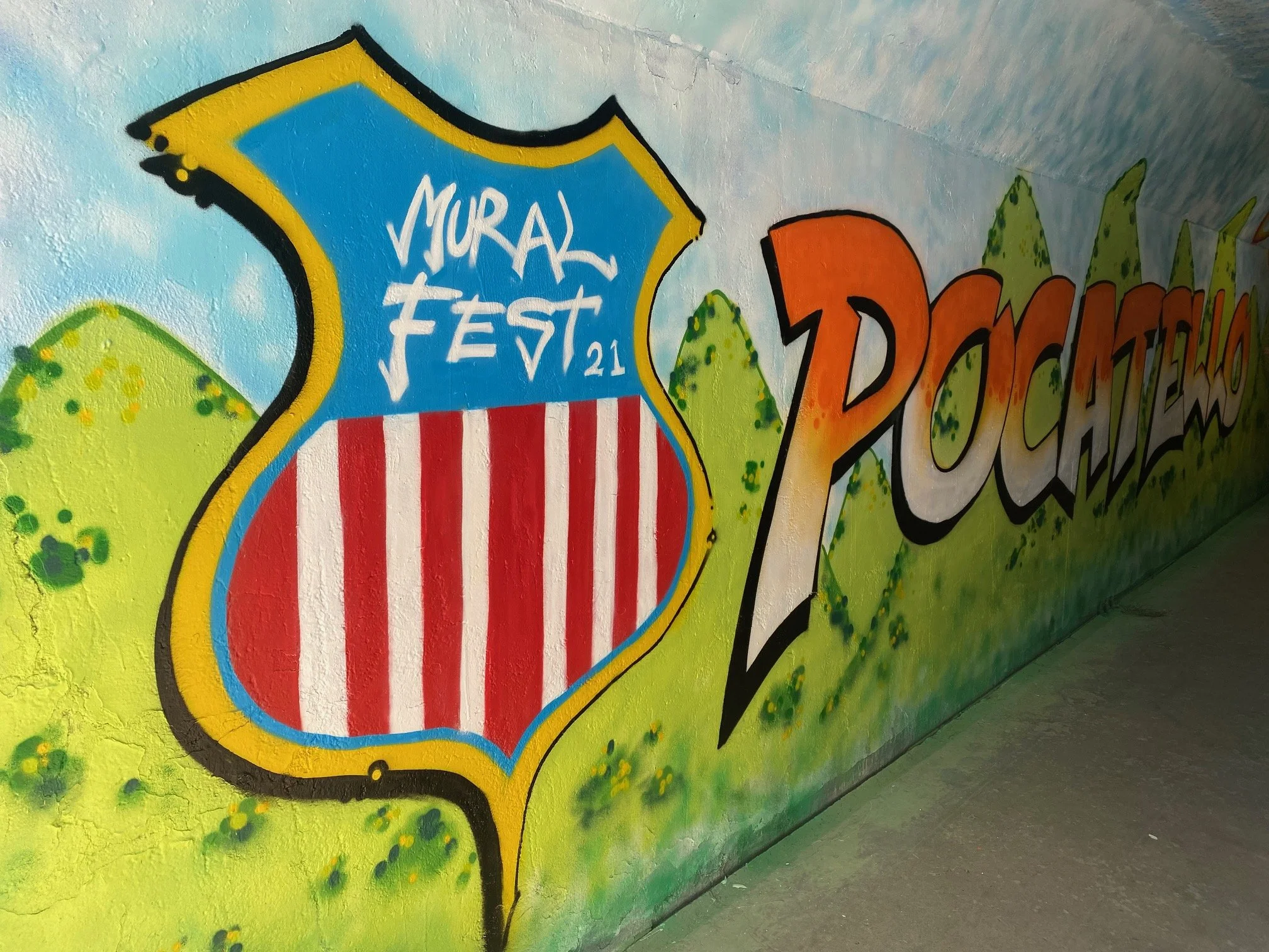 Colorful graffiti wall with a badge-shaped design that reads 'MURAL FEST 21'. The background has a bright yellow color with green accents, and a part of the adjacent graffiti says 'DREAM'.