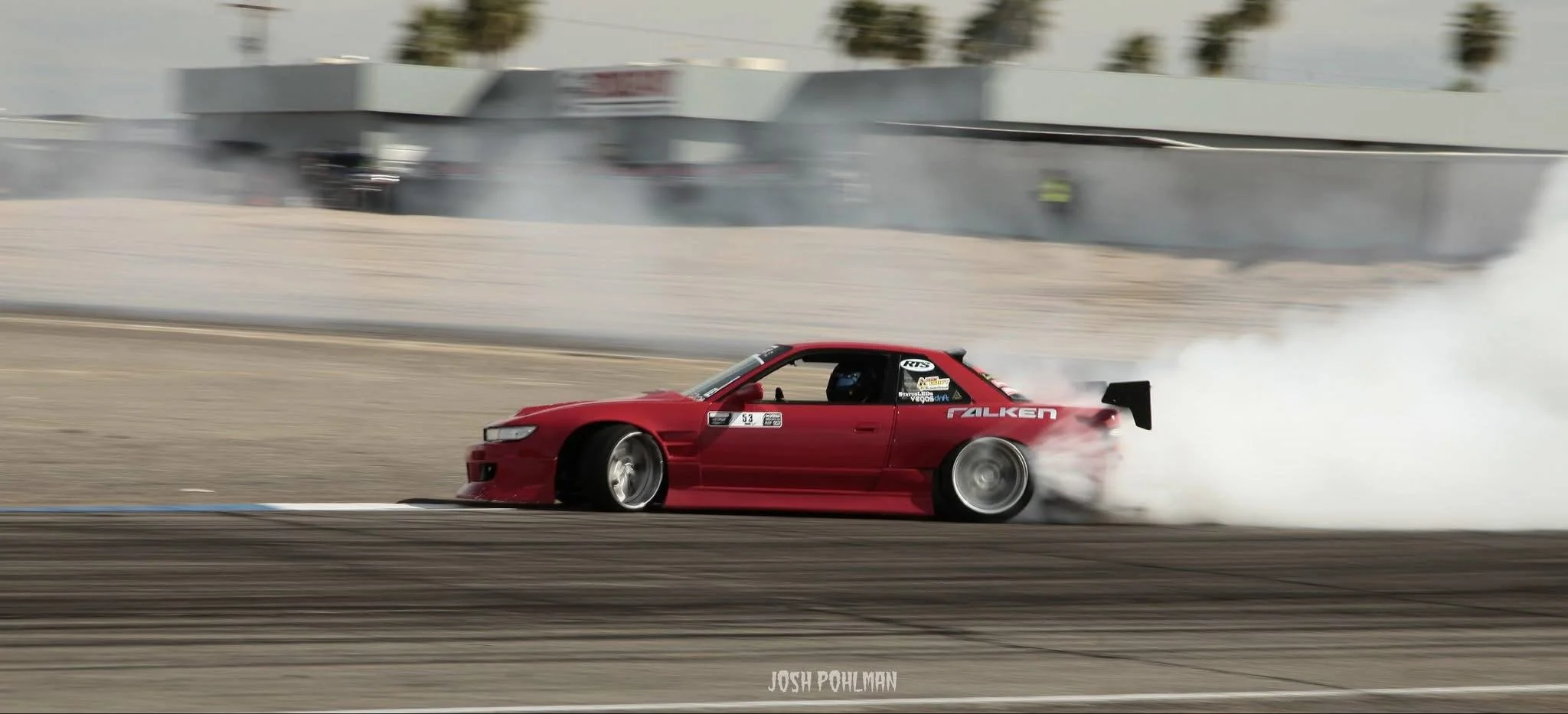 A red racing car drifting on a race track, producing smoke from its rear tires, with a blurred background of structures and palm trees.