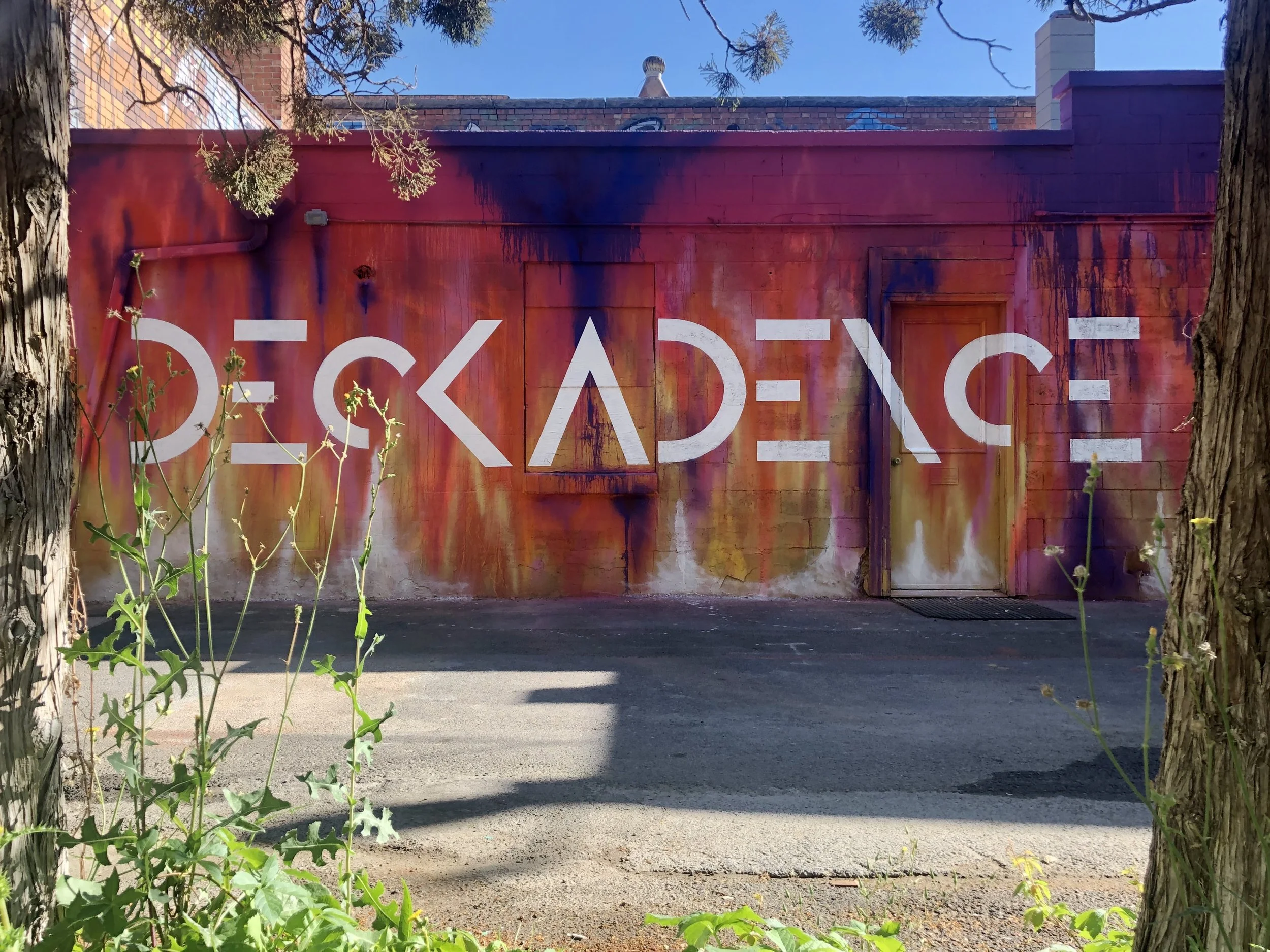 A vibrant paint dripped wall with white painted letters spelling out 'deckadence' , partially obscured by a tree trunk and green plants in the foreground. The wall has some paint streaks and a door on the right side.