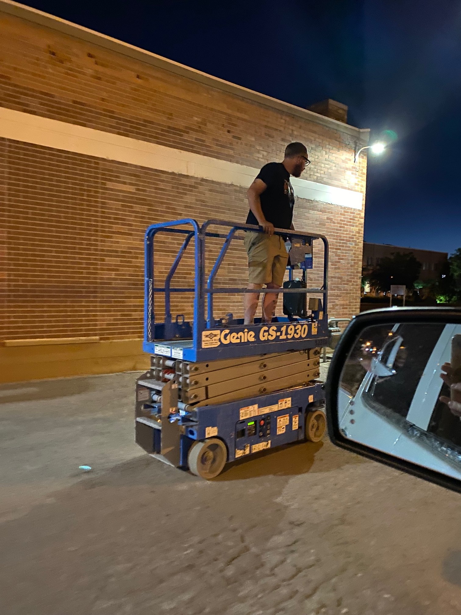 Night scene showing a person standing on a blue Genie GS-1930 lift platform attached to a vehicle, working on an exterior brick wall illuminated by a wall-mounted light.