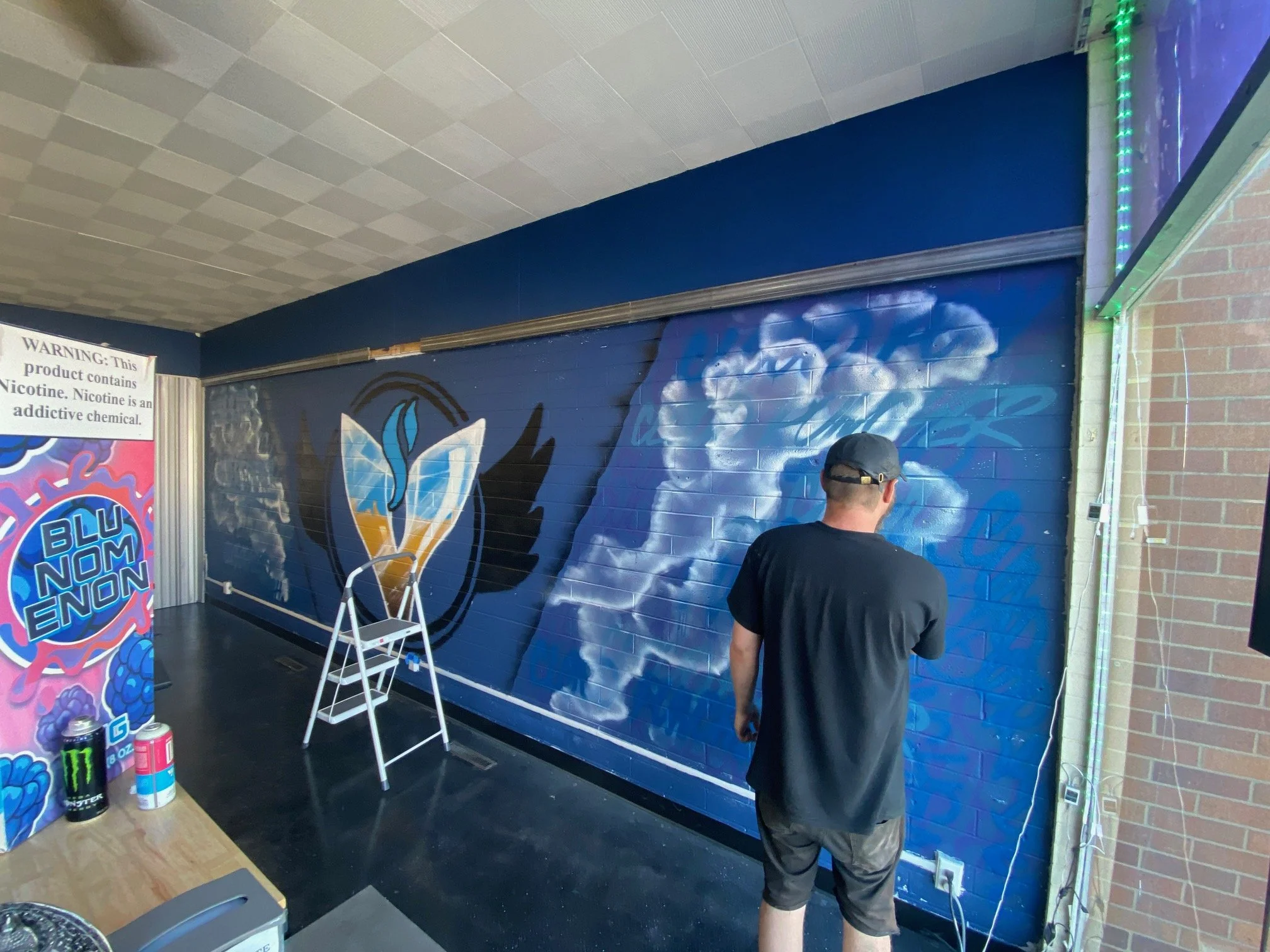 A person wearing a black cap and black shirt is standing outside the entrance of a building with a blue wall and mural artwork. Two spray paint cans and a ladder are placed on the ground nearby. Part of a colorful sign with the word 'BLUNON' is visib