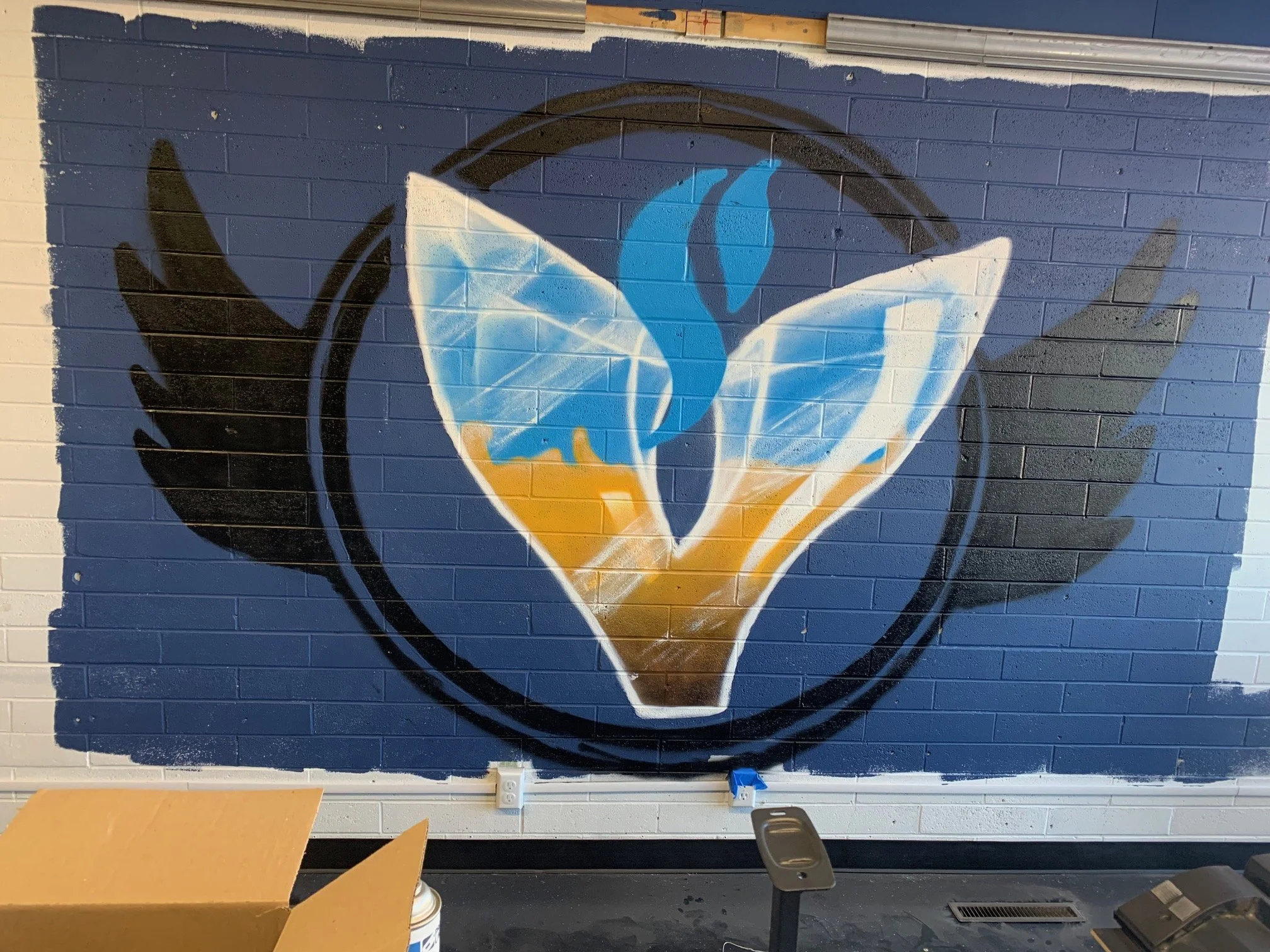 A painted mural of a steaming cup of coffee or tea on a brick wall, with a black circular border around it. The mural uses blue, white, black, and orange colors.