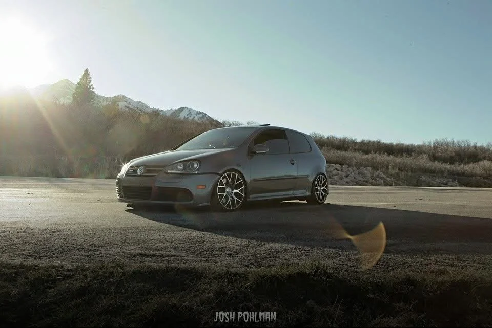 A gray Volkswagen Golf parked on a road with mountains and clear sky in the background during sunset or sunrise.